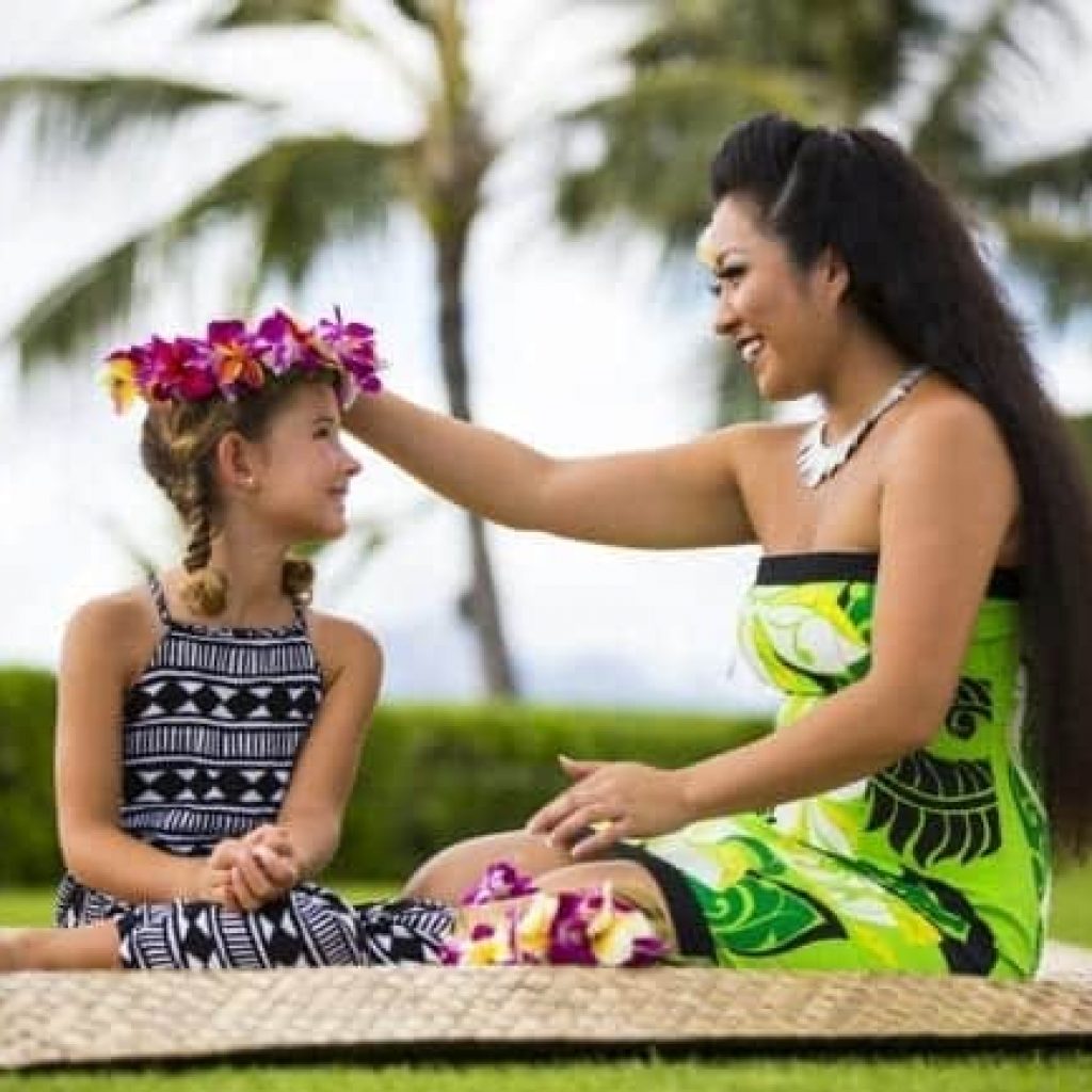 woman sitting on ground placing crown of flowers onto childs head at a hawaiian luau