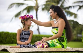 woman sitting on ground placing crown of flowers onto childs head at a hawaiian luau